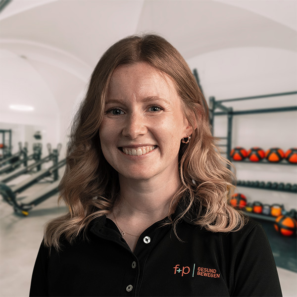 Marlene, Mitarbeiterin von f+p Gesund Bewegen, lächelt in der Trainingsstätte im Allgäu; im Hintergrund sind rote Gym-Bälle und Trainingsgeräte sichtbar.