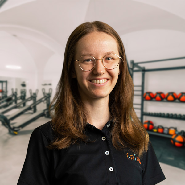 Michaela mit braunen Haaren und Brille trägt ein schwarzes Polohemd, im Hintergrund Trainingsgeräte und Hanteln – eine Szene aus dem f+p Gesundheitszentrum im Allgäu