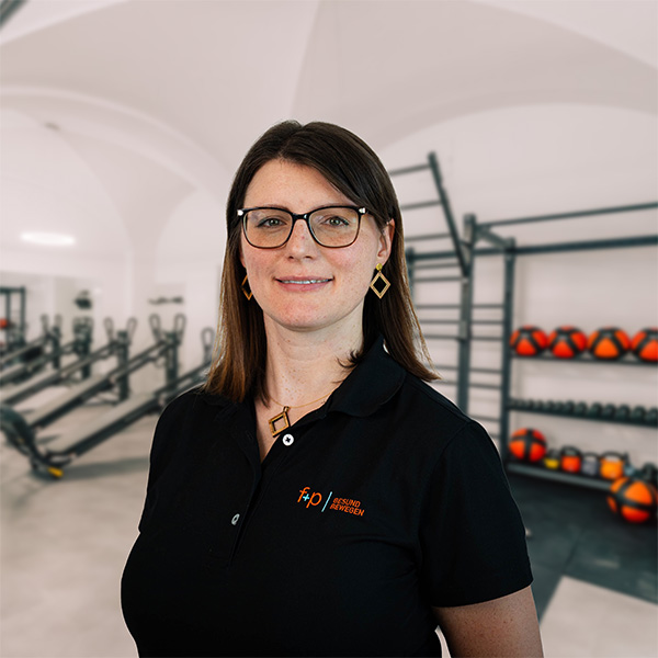 Frau mit Brille und schwarzem Poloshirt steht in einem Fitnessraum mit Trainingsgeräten.