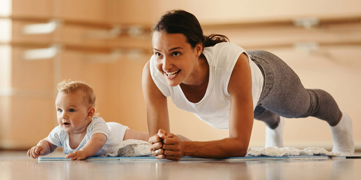 Eine Mutter führt eine Plank-Übung auf einer Yogamatte aus, während ihr Baby neben ihr liegt und lächelt.
