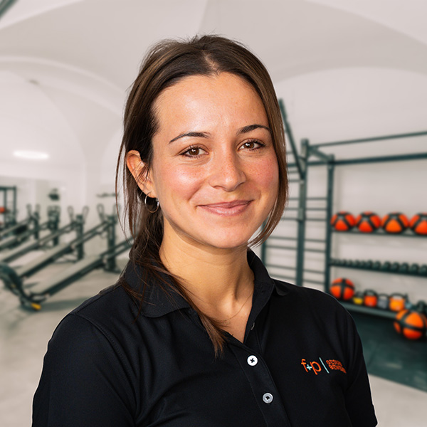 Frau mit dunklen Haaren und schwarzem Poloshirt in einem Fitnessstudio mit Trainingsgeräten im Hintergrund.