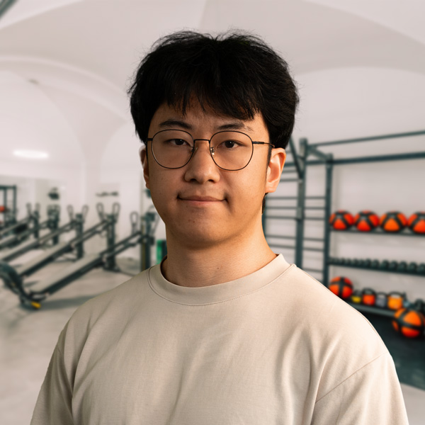 Yi_R, dunkle Haare und runde Brille, posiert im Fitnessstudio vor Trainingsgeräten und Kugelhanteln; klare Gesichtszüge, neutrale Mimik, Hintergrund mit Geräten