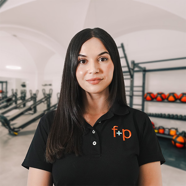 Zerina Suljic Frau mit langen, dunklen Haaren in einem schwarzen Polo-Shirt mit Logo in einem Fitnessstudio.