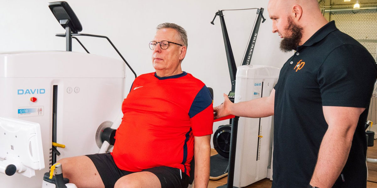 Mann in rotem Shirt sitzt an einem Fitnessgerät, während ein Trainer ihn anleitet.