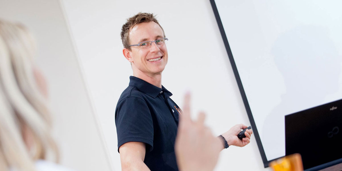 Mann mit Brille steht vor einer Präsentationsleinwand und hält einen Zeigestab in der Hand.