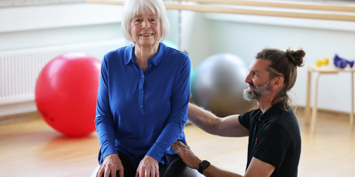 Ältere Frau sitzt auf einem Gymnastikball, während ein Physiotherapeut sie unterstützt.