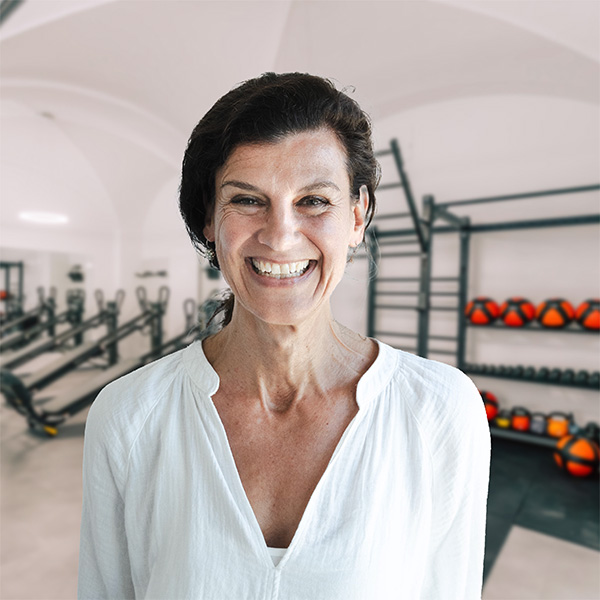 Daniela, Fachärztin für Allgemeinmedizin und Psychotherapie, lächelt in einem weißen Hemd im Fitnessstudio Allgäu; im Hintergrund Trainingsgeräte und Gym-Atmosphäre.
