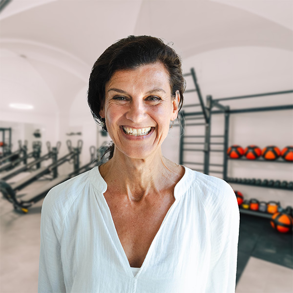Lächelnde Frau mit dunklen Haaren in einem Fitnessstudio mit Trainingsgeräten im Hintergrund.