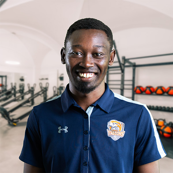 Frankline, dunkelhäutiger Mann mit kurzem Haarschnitt, lächelt in einem Fitnessstudio. Er trägt blaues Polo mit dem Falcons-Logo; der Hintergrund zeigt Trainingsgeräte.