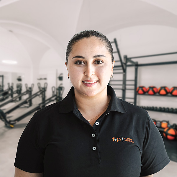 Ilhem in einem dunklen Polohemd der Praxis f+p Gesund Bewegen, Hintergrund mit Fitnessgeräten und Trainingsequipment, klare Ausstrahlung, moderne Räume.