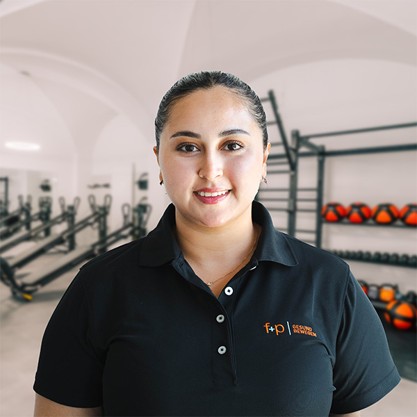 Ilhem Saussia Porträt einer Frau in einem schwarzen Poloshirt mit Logo vor Fitnessgeräten im Hintergrund.