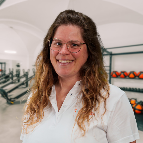 Jessica, Mitarbeiterin bei f+p Gesund Bewegen im Allgäu, lächelt in einem Fitnessstudio. Sie trägt eine Brille, ihr langes braunes Haar fällt locker, sie trägt ein weißes Polohemd; im Hintergrund Trainingsgeräte und Hanteln.