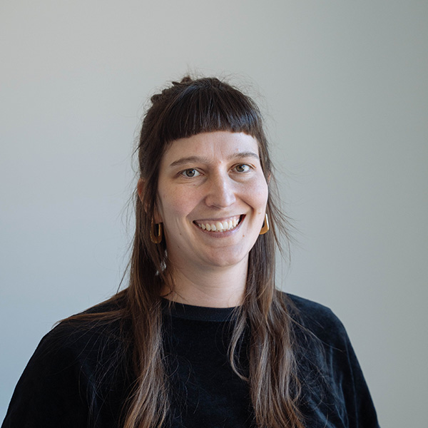 Katharina von f+p Gesund Bewegen mit braunen Haaren und goldenen Ohrringen, trägt schwarzes Oberteil, freundliches Lächeln, hellgrauer Hintergrund