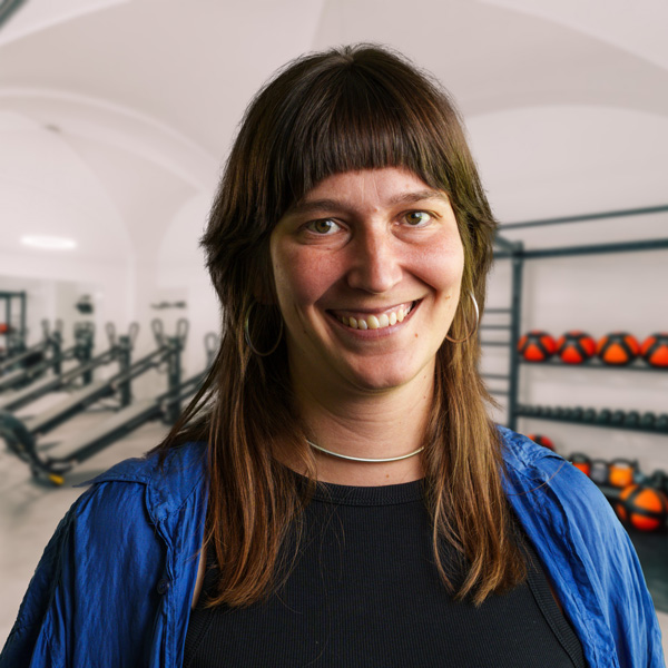 Frau mit langen Haaren und blauer Jacke lächelt in einem Fitnessstudio mit Fitnessgeräten im Hintergrund.