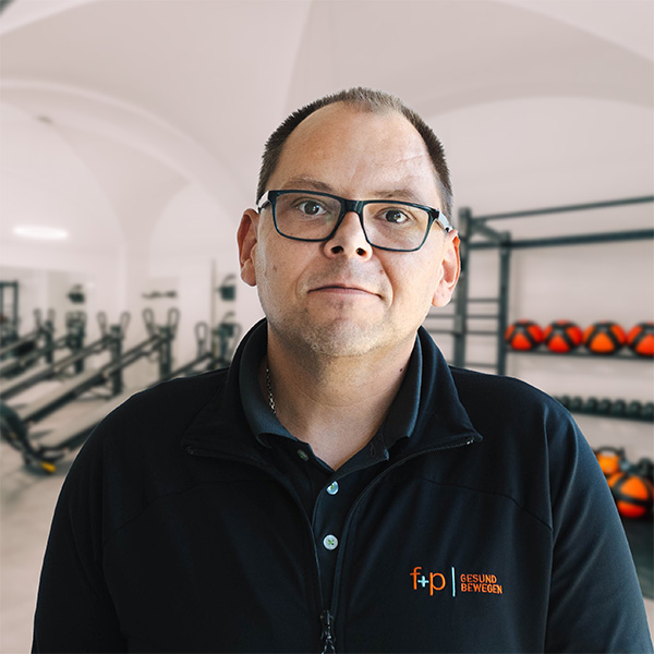 Kevin Fahrservice von f+p Gesund Bewegen trägt schwarze Jacke und Brille, steht im hellen Fitnessstudio, im Hintergrund sind Trainingsgeräte und rote Medizinbälle sichtbar