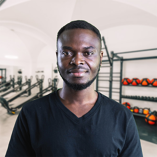 Mark – Mitarbeiter von f+p Gesund Bewegen im Allgäu, portraitierter Mann mit dunkler Hautfarbe, schwarzes V-Neck T-Shirt, sportive Gym-Umgebung hinter ihm