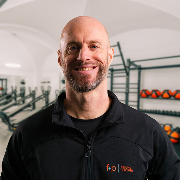 Mann mit Glatze und Bart in einem schwarzen Trainingsanzug, lächelt in einem Fitnessstudio.