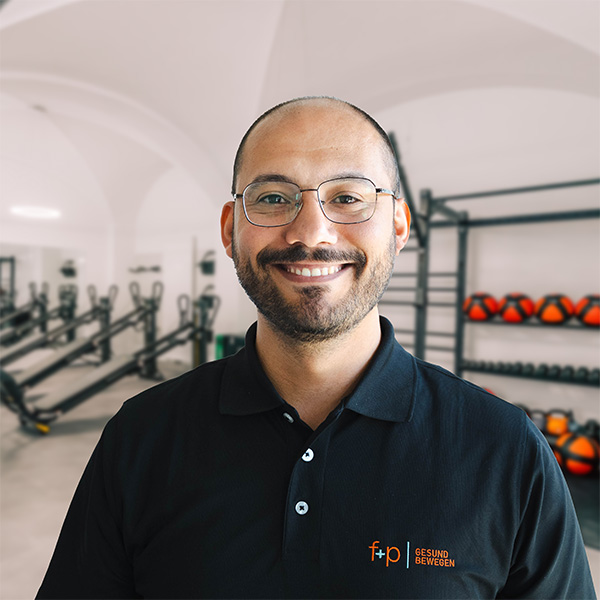 Wajdi, Mitarbeiter von f+p Gesund Bewegen im Allgäu, trägt dunkles Polohemd und Brille vor einem Fitnessraum mit Trainingsgeräten und Hanteln.