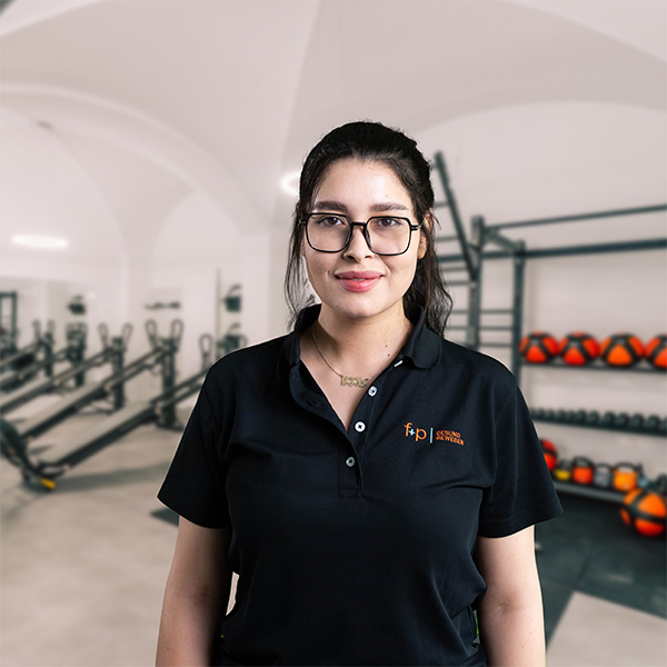 Frau mit Brille in einem schwarzen Polo-Shirt vor einem Fitnessstudio mit Geräten im Hintergrund.