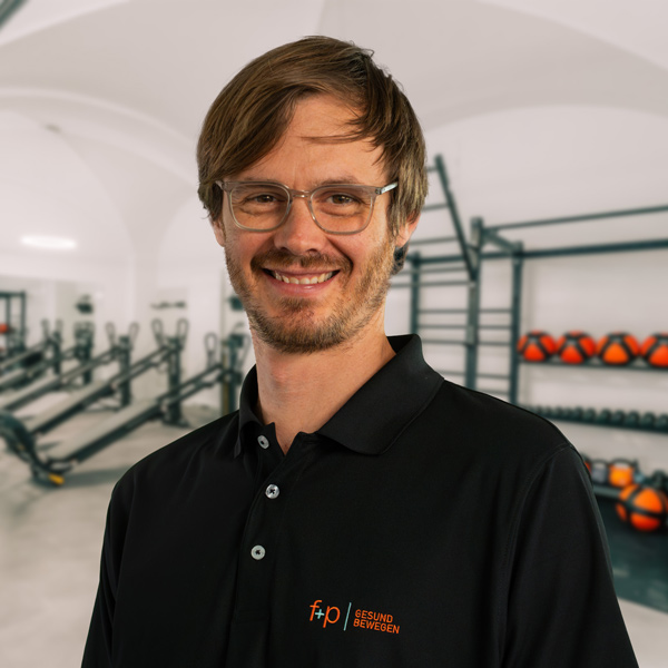 Yannick mit Brille und braunen Haaren trägt schwarzes Polo mit f+p Logo und steht lächelnd in einem hellen Trainingsraum der Allgäu-Einrichtung von f+p Gesund Bewegen