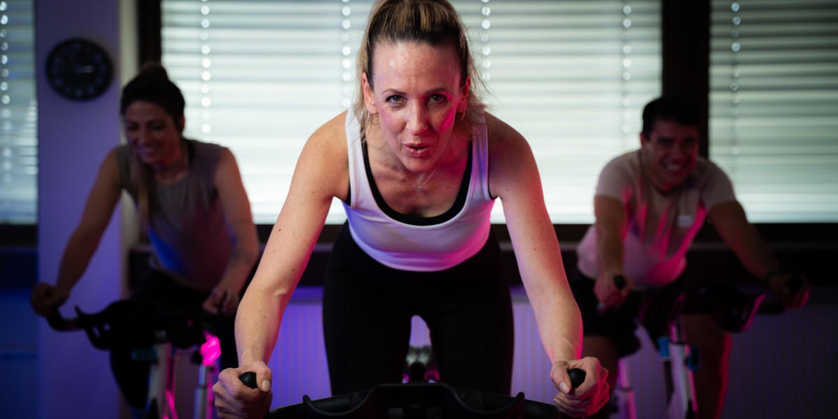 Drei Personen nehmen an einem Indoor-Cycling-Kurs teil, eine Person ist im Vordergrund aktiv auf dem Fahrrad.