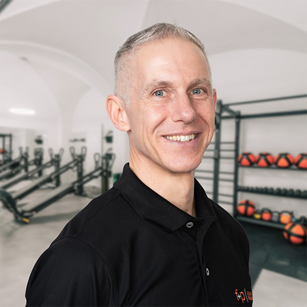 Christian – Therapeutische Leitung bei f+p Gesund Bewegen im Allgäu, lächelnd vor einem modernen Therapieraum mit Trainingsgeräten im Hintergrund, graue Haare und schwarze Polo-Bluse.