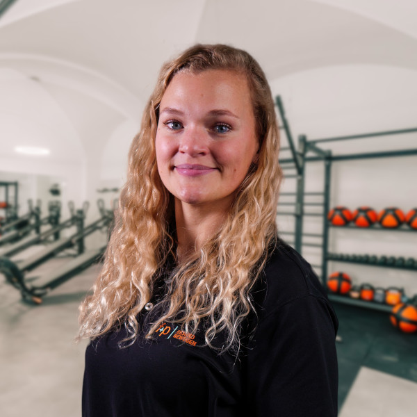 Eine Person mit langen, lockigen Haaren trägt ein schwarzes Polohemd und steht in einem Fitnessstudio.