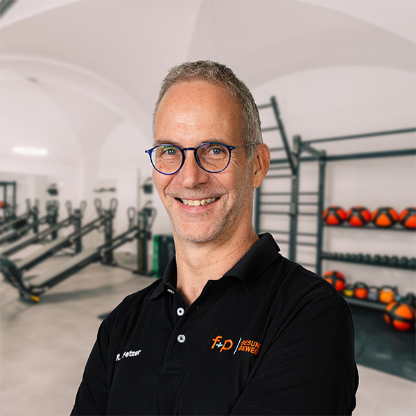 Mann mit Brille und schwarzem Polo-Shirt in einem Fitnessstudio mit Trainingsgeräten im Hintergrund.