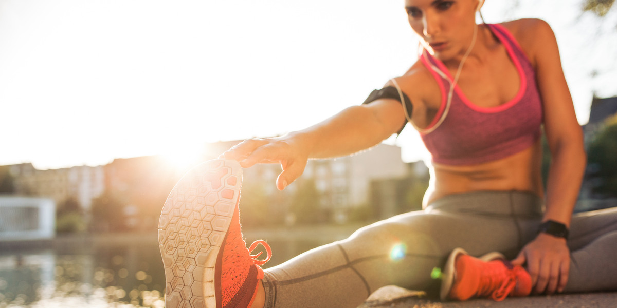 Eine Frau in Sportkleidung dehnt ihre Beine im Freien, während die Sonne im Hintergrund scheint.