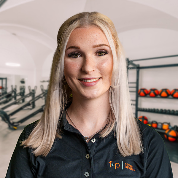 Annika, Mitarbeiterin von f+p Gesund Bewegen im Allgäu, blondes Haar, freundliches Lächeln, schwarze Bluse mit Logo, Hintergrund Trainingsraum mit Geräten.