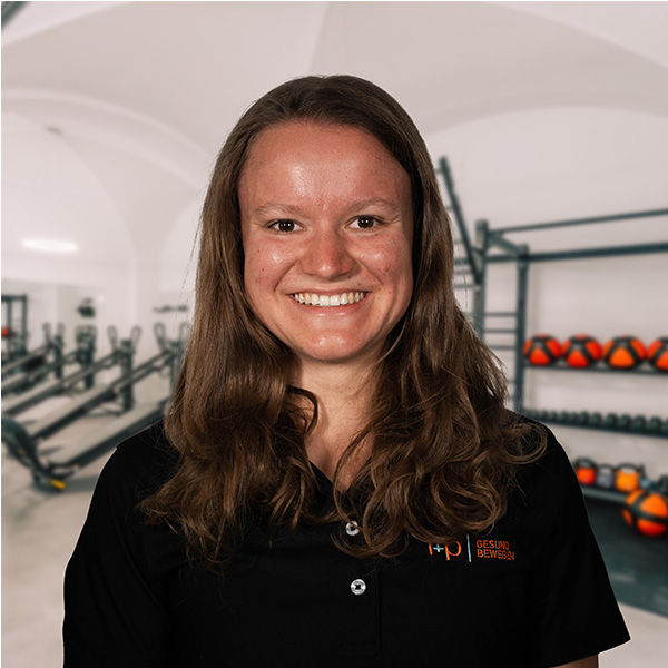 Frau mit langen Haaren, die ein schwarzes Polohemd trägt, vor einem Fitnessstudio-Hintergrund.