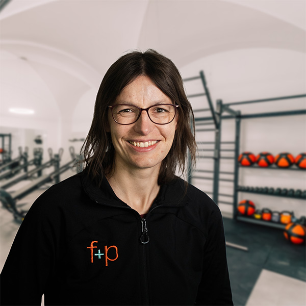 Frau mit Brille und schwarzem Trainingsanzug lächelt in einem Fitnessstudio.