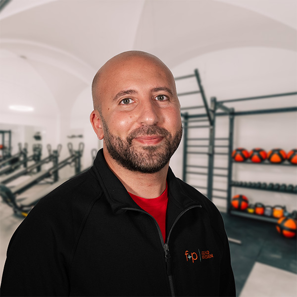 Hamdi, glatzköpfiger Physiotherapeut von f+p Gesund Bewegen im Allgäu, trägt schwarze Jacke und rote Shirt, lächelt, Trainingsraum mit Regalen und Medizinball-Übungsequipment im Hintergrund