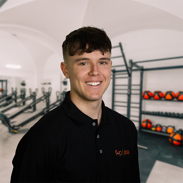 Junger Mann mit kurzem Haar und lächelndem Gesicht in einem Fitnessstudio mit Trainingsgeräten im Hintergrund.
