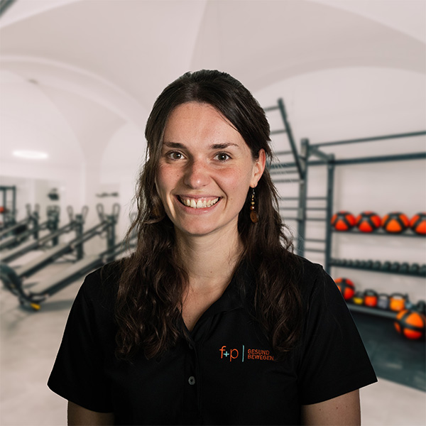 Frau mit langen, dunklen Haaren und schwarzem Hemd, lächelt in einem Fitnessstudio mit Trainingsgeräten im Hintergrund.