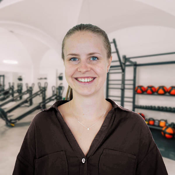 SaBi_web Lächelnde Frau mit blonden Haaren in einem Fitnessstudio mit Trainingsgeräten im Hintergrund.