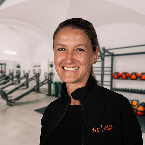 Stephanie in schwarzer Sportjacke mit Logo f+p Gesund Bewegen im Trainingsraum, Allgäu, Hintergrund mit Trainingsgeräten und Hanteln, freundliches Lächeln
