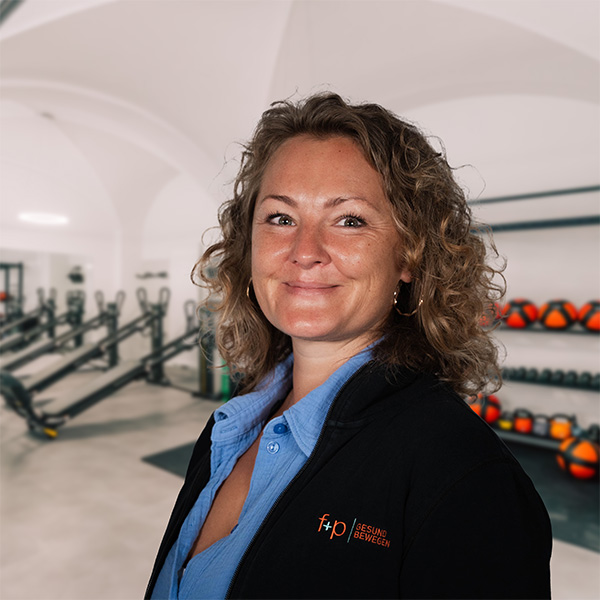 Frau mit lockigem Haar, die in einer Sporteinrichtung steht, mit Fitnessgeräten im Hintergrund.