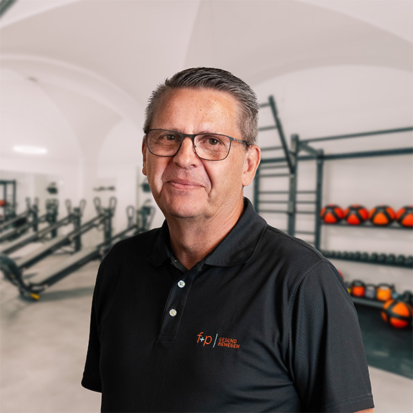 Mann mit Brille und schwarzem Poloshirt steht in einem Fitnessstudio mit Trainingsgeräten im Hintergrund.