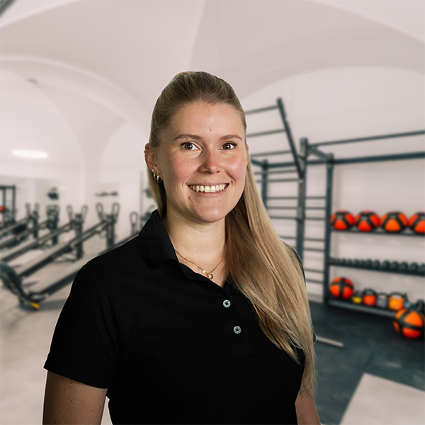 Frau mit langen Haaren in einem schwarzen Poloshirt steht in einem Fitnessstudio mit Trainingsgeräten im Hintergrund.
