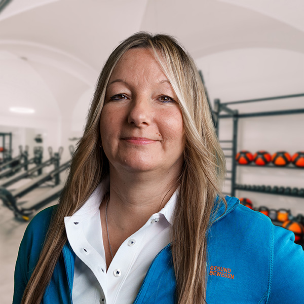 Frau mit langen, blonden Haaren, die eine blaue Jacke trägt und in einem Fitnessraum steht.