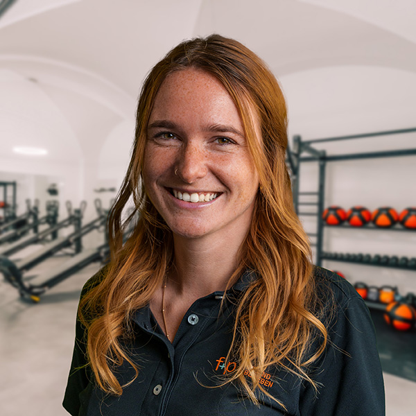 DésiréeGr Frau mit langen, roten Haaren und einem freundlichen Lächeln in einem Fitnessstudio mit Trainingsgeräten im Hintergrund.