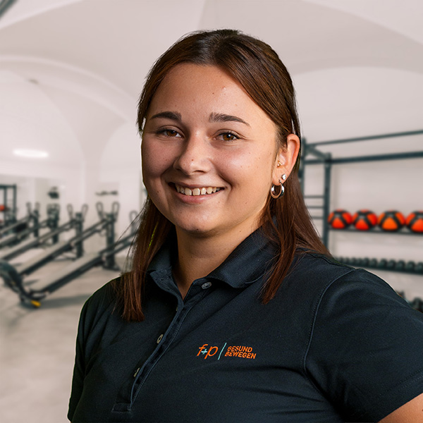 JessicaFo Lächelnde Bewegungsexpertin in einem Sportstudio mit Fitnessgeräten im Hintergrund.