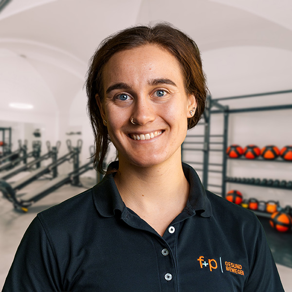Trainerin mit kurzen braunen Haaren und einem schwarzen Poloshirt in einem Fitnessstudio mit Trainingsgeräten im Hintergrund.