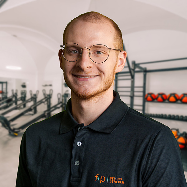 Mann mit Brille trägt ein schwarzes Poloshirt und lächelt in einem Fitnessstudio mit Trainingsgeräten im Hintergrund.