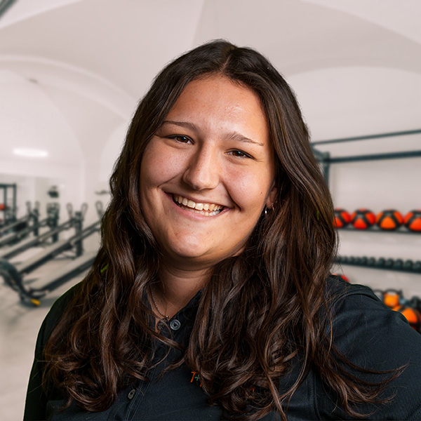 TamaraGa Lächelnde Frau mit langen, lockigen Haaren, die in einem Fitnessstudio steht, mit Fitnessgeräten im Hintergrund.