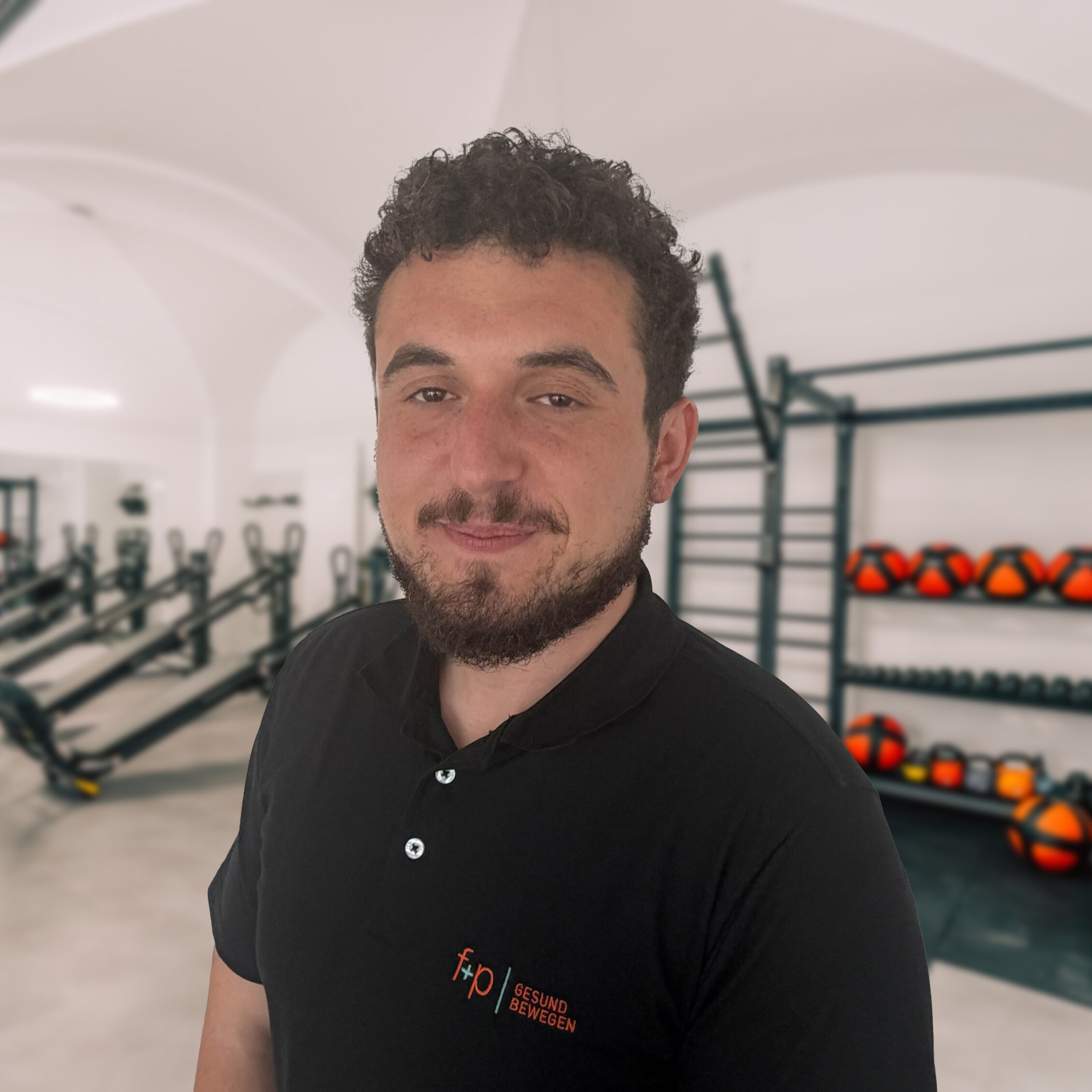 Tunahan mit lockigem Haar und schwarzem Shirt im Trainingsraum von f+p Gesund Bewegen, Physiotherapie mit Übungsgeräten im Hintergrund.