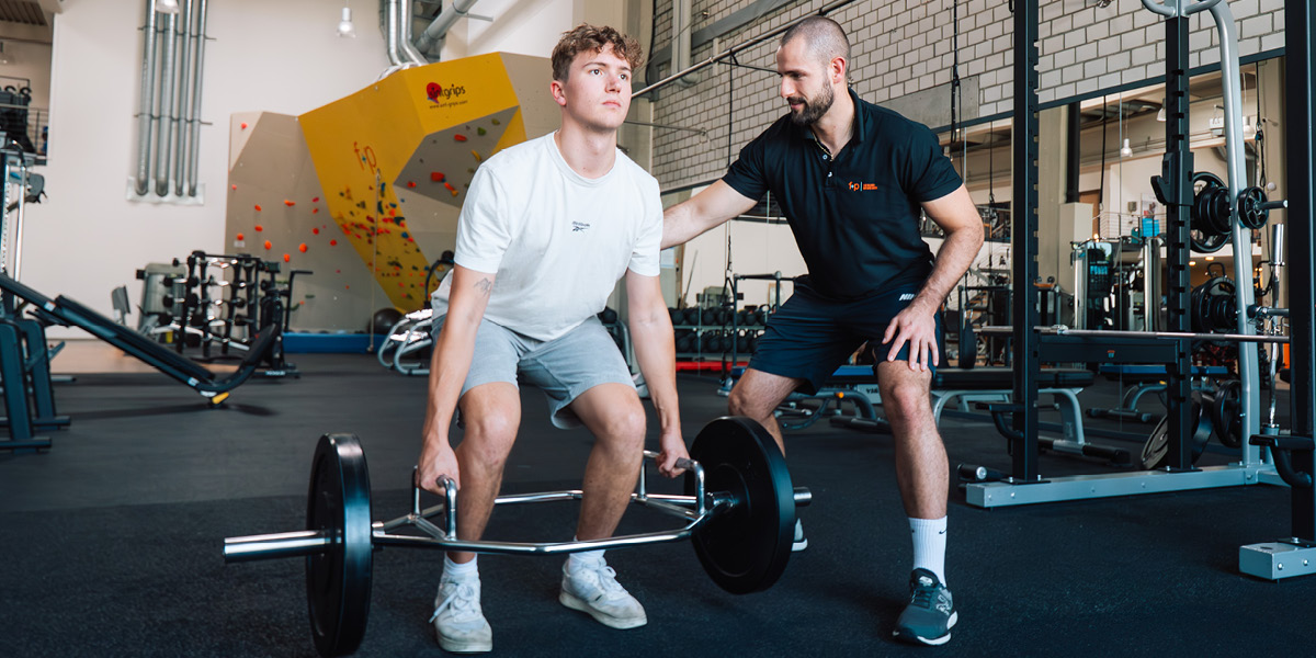 Junger Mann hebt eine Langhantel mit Unterstützung eines Trainers in einem Fitnessstudio.