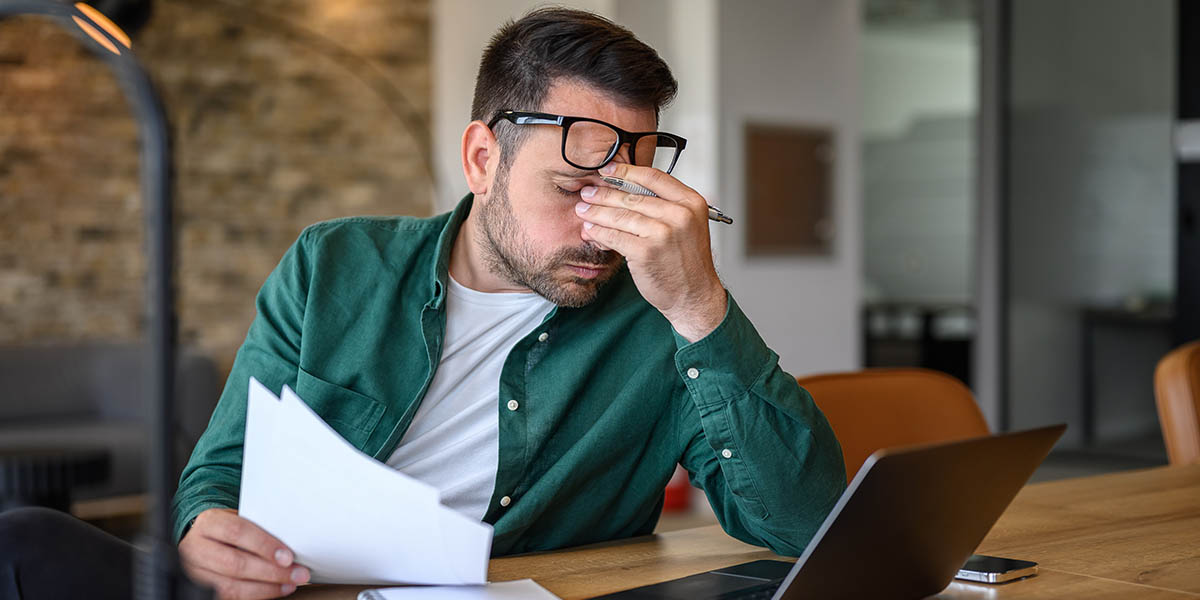 Mann mit Brille, der frustriert auf seine Unterlagen schaut und eine Hand an die Stirn legt.