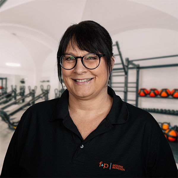 Frau mit Brille und schwarzem Polohemd steht in einem Fitnessraum mit Trainingsgeräten im Hintergrund.
